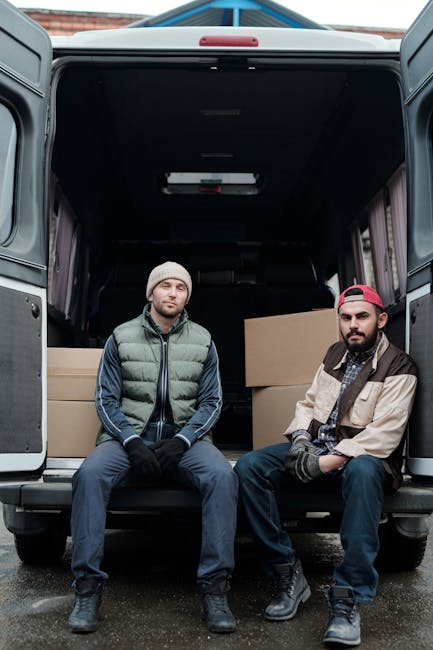 A man wearing a cap, dark jacket, and trousers is engaged in packing or preparing for a home relocation outside a modern building with large glass panels. He is standing next to several cardboard boxe